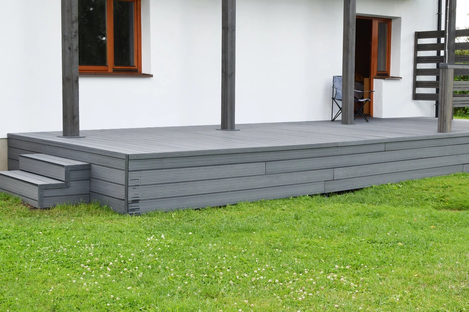 Partie d'une maison résidentielle blanche avec portes brunes, fenêtres et terrasse en matériaux composites gris foncé ou anthracite wpc avec balustrades en bois à l'extérieur